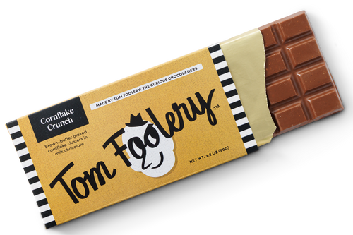 A single wrapped chocolate bar on a clean white background. The wrapper is tan with black-and-white striped edges, large 'Tom Foolery' script, and 'Cornflake Crunch' text.