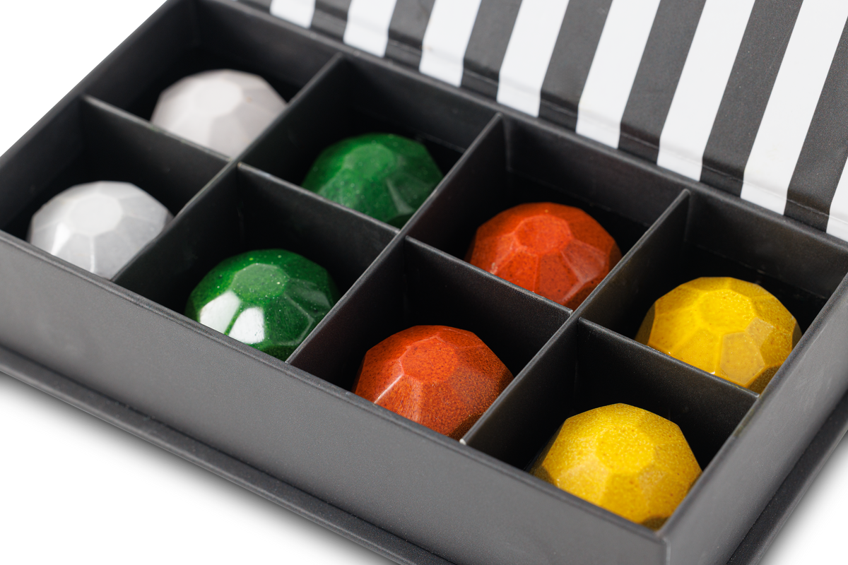 A close-up of a black compartmentalized box containing faceted gem-like pieces in white, green, orange-red, and yellow, with a black-and-white striped lid visible in the background.